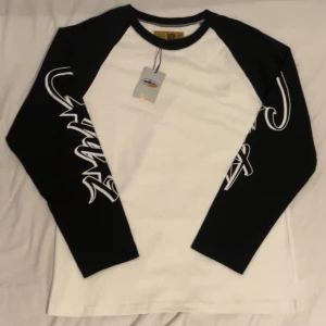 Svartvit långärmad Cortez Guerillaz tröja - Säljer en svartvit långärmad tröja från Cortez Guerillaz i storlek S. Tröjan har vita ärmar med svart grafiskt tryck och svart krage. Materialet är mjuk bomull och passformen är normal. Perfekt för dig som gillar streetwear och snygga detaljer.