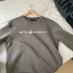 Grå Sail Racing sweatshirt M - Snygg frostgrå sweatshirt från Sail Racing i storlek M. Tröjan har rund hals, lång ärm och vit logotyp tryckt på bröstet. Materialet är mjuk bomull med insida i fleece för extra komfort. Perfekt för chill dagar och enkel att matcha med jeans eller joggers. 