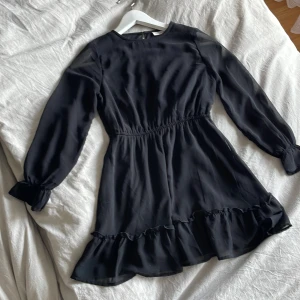 Svart volangklänning från Kappahl - Supersnygg svart klänning från Kappahl med volangdetaljer längst ner och vid ärmsluten. Den har långärmade ärmar och en markerad. Bra skick🎀