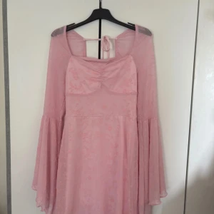 Rosa kortklänning med vida ärmar från SHEIN - Supersöt ljusrosa kortklänning från SHEIN med vida, transparenta ärmar och knytning i ryggen. Klänningen har ett diskret blommigt mönster och lätt rynkning vid bysten. Perfekt för dig som gillar romantisk och feminin stil.