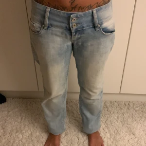 Ljusblå jeans med dubbelknäppning - Säljer ett par ljusblå jeans med cool tvättad look och dubbelknäppning fram. Jeansen har fem fickor, låg midja och raka ben. Materialet är klassisk denim och passformen är normal. Perfekt för dig som gillar avslappnad stil och snygga detaljer. Midjemått: 41cm