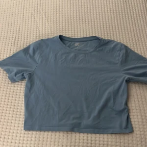 Blå croppad t-shirt från 157 - Snygg ljusblå croppad t-shirt från 157 i storlek M. T-shirten har rund hals och korta ärmar, tillverkad i mjuk bomull med lite stretch för extra komfort. Perfekt för dig som gillar enkel och clean stil. Tröjan är en magtröja och jag skulle säga att den är mycket mindre än vad storleken säger