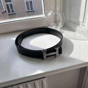 Svart skärp med H-spänne - Snyggt svart skärp med ett stort H-format spänne i silverfärgad metall. Bältet är gjort av skinn med vit kontrastsöm längs kanterna och har en klassisk, stilren look som verkligen sticker ut. Perfekt för dig som gillar exklusiva accessoarer.