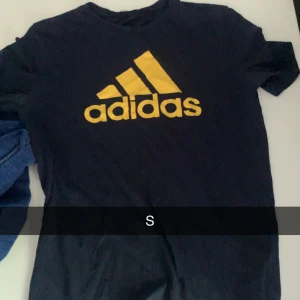 Svart Adidas t-shirt med gul logga - Svart t-shirt från Adidas med stor gul logga på bröstet. Klassisk rund hals och korta ärmar. Perfekt för dig som gillar sportiga och stilrena plagg. Materialet känns mjukt och skönt mot huden.