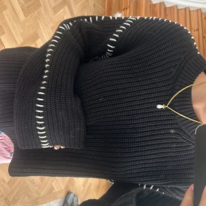 Svart stickad tröja med vita detaljer - Säljer en svart stickad tröja med grov ribbad struktur och vita dekorativa sömmar längs ärmar och axlar. Tröjan har lång ärm och en relaxed passform, perfekt för dig som gillar en chill och stilren look. Superenkel att matcha med jeans eller cargo. Den säljs inte längre på Gina tricot