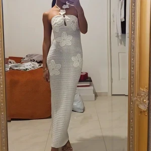 Vit lång virkad klänning med blommor - Supersnygg långklänning i vit virkad stil med stora blommor som detaljer. Klänningen är axelbandslös med halterneck och har en tight passform som framhäver figuren. Perfekt för sommarens alla tillfällen, med unik och trendig look.