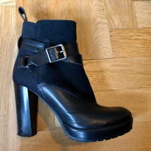 Svarta stövletter från G-Star Raw - Snygga svarta stövletter från G-Star Raw med chunky hög klack och plattform. Skorna har coola spänndetaljer och är tillverkade i en mix av skinn och mocka. Perfekt för dig som vill ha en edgy look med italiensk kvalitet och grym passform.