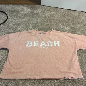 Croppad rosa frottétröja BEACH please - Säljer en croppad rosa frottétröja med trycket 'BEACH please' i vitt på bröstet. Tröjan har korta ärmar och en loose passform, perfekt för chill dagar eller strandhäng. Mjuk och skön känsla tack vare frottématerialet.