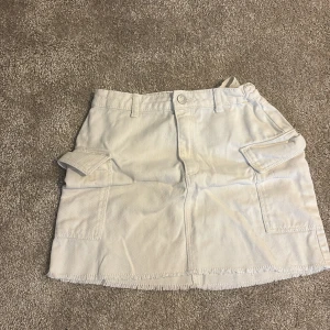 Vit kortkjol med fickor från H&M - Snygg vit kortkjol från H&M med fransad kant och stora fickor på sidorna. Kjolen har bälteshällor och knappstängning fram. Tillverkad i 100% bomull för en skön och avslappnad känsla. Perfekt för dig som gillar street och utility-vibe.