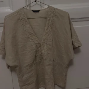 Beige blus med v-ringning från Zara - Säljer en beige blus från Zara i storlek XS. Blusen har en djup v-ringning med snygga broderade detaljer längs halsen och ärmarna. Tillverkad i mjukt linne, med korta ärmar och avslappnad passform. Perfekt för dig som gillar en chill och stilren look. Aldrig använd säljer pågrund av att jag använder inte priset är billigt för äkta linne tyg!