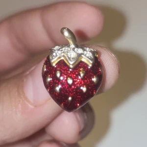 Glittrigt jordgubbshänge i guld och rött - Unikt hänge format som en jordgubbe med glittrig röd yta och guldfärgade detaljer. Toppen är dekorerad med små klara stenar som ger extra bling. Perfekt som charm till halsband eller armband för dig som gillar färgglada och söta smycken.