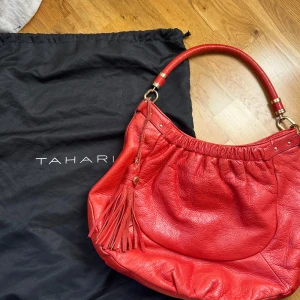 Röd axelväska från Tahari i skinn - Snygg röd/orange/korall axelväska från Tahari i mjukt skinn med en bred axelrem och coola gulddetaljer. Väskan har en stor tofs som hänger på sidan och ett rymligt innerfack med dragkedja. Perfekt för dig som vill ha en färgstark accessoar med plats för allt du behöver.