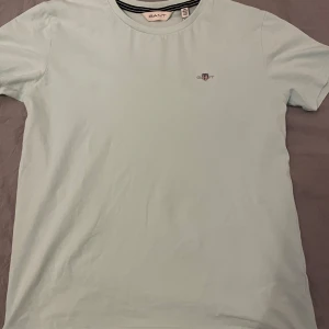 Turkos GANT t-shirt med logga - Fräsch Turkus t-shirt från GANT med klassisk rund hals och broderad logga på bröstet. T-shirten är enkel och stilren, perfekt för dig som gillar cleana plagg. Materialet är mjuk bomull som känns skön mot huden. Passar dig som vill ha en chill och snygg look.