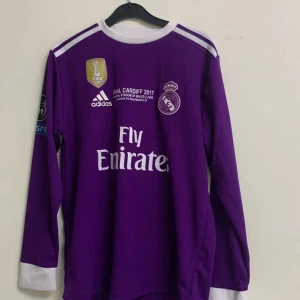 Real Madrid Ronaldo 7 lila långärmad tröja - Champions League Final Cardiff 2017 Real Madrid tröja i lila med vita detaljer, långärmad och med tryck 'Ronaldo 7' på ryggen. Adidas-logga, FIFA-märke och Fly Emirates sponsor på bröstet. Materialet är lätt och andas, perfekt för fotboll eller samling.