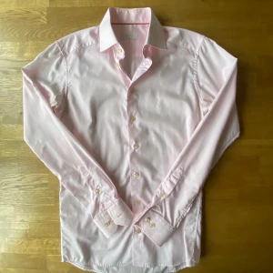 Rosa randig skjorta från Eton  - Snygg rosa skjorta från Eton i slim fit med diskreta vita ränder. Klassisk krage, långa ärmar och pärlemorknappar framtill. Tillverkad i mjuk bomull som känns skön mot huden. Perfekt för dig som gillar stilrena detaljer och en fräsch look.