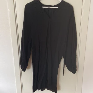 Svart omlottklänning från Lindex M - Svart omlottklänning från Lindex i storlek M. Klänningen har lång ärm, v-ringad hals och knytband i midjan. Materialet är lätt och har en skrynklig struktur som ger en cool och avslappnad vibe. Perfekt för dig som gillar enkel och stilren look.