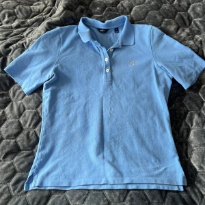 Blå GANT pikétröja i bomull - Snygg ljusblå pikétröja från GANT i storlek S. Klassisk krage, tre vita knappar framtill och diskret broderad logga på bröstet. Tillverkad i 100% bomull för skön och luftig känsla. Perfekt till jeans eller shorts när du vill ha en clean look.