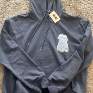 Mörkblå hoodie från Risky - Snygg mörkblå hoodie från Risky med stor huva och snörning. Framtill finns ett fluffigt ljusblått R-märke och på huvan är Risky tryckt i droppande stil. Perfekt för chill dagar och streetwear. Materialet känns mjukt och är troligen bomull. Helt ny