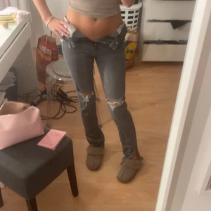 Grå ripped jeans med hög midja - Säljer ett par grå jeans med slitna detaljer vid knäna och låg midja. Modellen är straight med klassisk femficksdesign och knappstängning. Jeansen har en cool, avslappnad vibe och passar perfekt till sneakers eller boots.