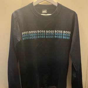 Svart långärmad tröja från BOSS - Snygg svart långärmad tröja från BOSS med rund hals och tryck i vitt och blått framtill. Tröjan har en klassisk passform och är gjord i mjuk bomull som känns skön mot huden. Perfekt för dig som gillar stilrena logotröjor med lite edge.