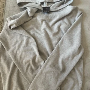 Grå Kashmir hoodie från Oscar Jacobsson storlek M - Snygg grå hoodie i mjukt bomullsmaterial från Oscar Jacobsson Tröjan har huva och långa ärmar, perfekt för chill dagar. Enkel och stilren design utan mönster eller detaljer, passar dig som gillar basic och comfy vibes.