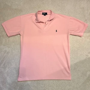 Rosa Ralph Lauren Piké - Snygg rosa pikétröja från Polo Ralph Lauren med klassisk krage och två knappar framtill. Tröjan har korta ärmar och den ikoniska broderade loggan på bröstet. Tillverkad i mjuk bomull som är skön mot huden. Perfekt för dig som gillar stilren och enkel design.