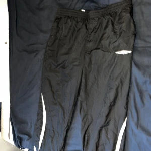 Svarta träningsbyxor från Umbro - Svarta träningsbyxor från Umbro med vita detaljer längs benen. Byxorna har resår i midjan och dragkedja vid bensluten . Lätta och luftiga, perfekta för sport eller chill. Klassisk sportig look. Riktiga från 2000 talet