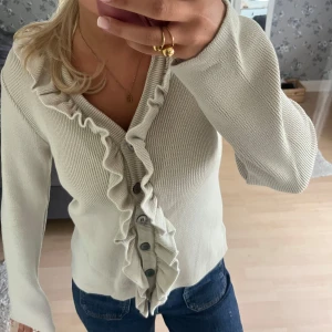 Beige kofta med volang och knappar - Supersöt beige kofta med volang längs v-ringningen och knappar framtill. Stickad i mjukt material och har långärmade ärmar. Perfekt att styla med jeans för en chill och snygg look. Volangdetaljen ger en extra touch av trend.