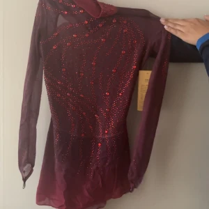 Vinröd glittrig konståkningsklänning med öppen rygg - Supersnygg vinröd konståkningsklänning med glittrande röda stenar och meshdetaljer. Klänningen har långärmade ärmar, hög krage och en öppen rygg.