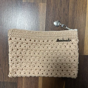 Beige stickad kuvertväska med hjärtdragkedja - Snygg beige kuvertväska som är stickad med ett unikt mönster. Väskan har en silvrig dragkedja med ett hjärta som detalj och en liten metallskylt med texten 'Handmade'. Perfekt för att bära småsaker och ge din outfit en personlig touch.