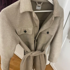 Beige overshirt från Zara med bälte/kappa - Snygg beige overshirt från Zara med klassisk krage, två bröstfickor med knappar och ett matchande bälte i midjan. Jackan har långa ärmar och svarta knappar framtill. Perfekt för lager-på-lager och chill stil under höst och vår.