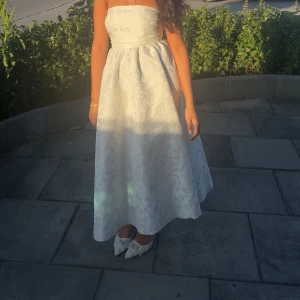 Vit axelbandslös långklänning - Säljer en elegant vit axelbandslös långklänning med vacker spetsstruktur och markerad midja. Klänningen har en utsvängd kjol som ger en snygg siluett och passar perfekt för student eller bal. Materialet är mjukt och har en lyxig känsla.