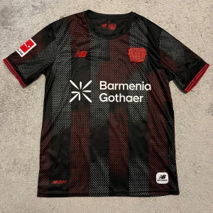 Bayer Leverkusen svart/röd matchtröja - Säljer en Bayer Leverkusen fotbollströja från New Balance i svart med röda och gråa prickiga ränder. Tröjan har klubbmärke och sponsortryck på bröstet, Bundesliga-märke på ärmen och röda detaljer vid ärmslut. Tillverkad i lätt och ventilerande funktionsmaterial.