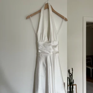 Vit kortklänning i satin med rosett - Supersnygg vit kortklänning från Zara i glansigt satinmaterial. Klänningen har halterneck, djup v-ringning och öppen rygg med en stor rosett bak. Denna har slutats säljas på Zara 💕 helt ny aldrig använd och prislappen är kvar!