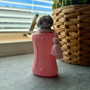 Parfym Delina Exclusif rosa flaska - Säljer en lyxig Delina Exclusif parfym från Parfums de Marly i en matt rosa flaska med dekorativa reliefdetaljer och en silvrig rund kork. Flaskan har en rosa tofs med snöre som ger en elegant vibe. Perfekt för dig som  gillar exklusiva dofter och snygg design. Endast testad 