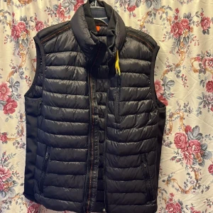 Svart dunväst från Parajumpers L - Snygg svart dunväst från Parajumpers med quiltad design och hög krage. Västen har dragkedja framtill, två sidofickor med zip och en gul detalj vid halsen. Insidan har fleecepaneler för extra komfort. Perfekt att slänga över hoodien när det är kyligt ute.