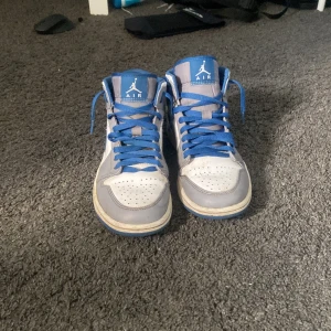 Nike Air Jordan 1 Mid blå/vit EUR 40 - Nike Air Jordan 1 Mid sneakers i blått och vitt med klassisk Jordan-logga på sidan och tungan. Skorna har blå snörning, perforerad tå och en snygg kontrast mellan de vita och blå panelerna. Perfekt för dig som gillar streetstyle och vill sticka ut med ikoniska sneakers. 
