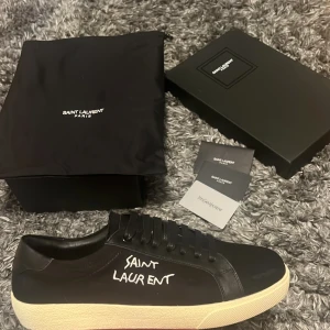 Svarta Saint Laurent sneakers - Svarta sneakers från Saint Laurent med vit text på sidan och guldfärgad logga på plösen. Skorna har snörning, rund tå och en vit platt sula med Saint Laurent Paris präglat undertill. Tillverkade i skinn med stilren design och medföljande originalkartong och dustbag.