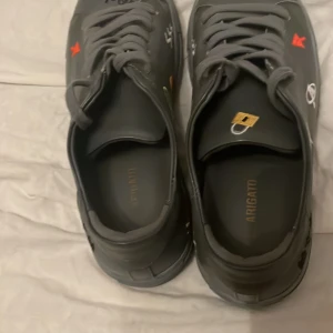 Grå sneakers från Axel Arigato - Säljer ett par grå sneakers från Axel Arigato med coola broderade detaljer i färg, som piller, gem och spruta. Skorna har snörning och platt sula, med en rund tå. Materialet är skinn och de har en unik streetstil som sticker ut.