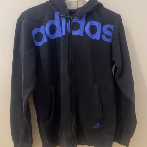 Svart hoodie från Adidas med blå logga - Svart hoodie från Adidas med stor blå Adidas-logga över bröstet. Tröjan har huva, dragkedja framtill och två fickor. 