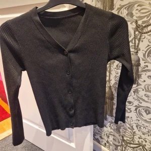 Svart ribbad kofta med v-ringning - Säljer en svart ribbad kofta med v-ringning och knappar framtill. Koftan har långa ärmar och är tillverkad i ett mjukt stickat material som sitter snyggt på kroppen. Perfekt att slänga över en t-shirt eller linne för en chill look.