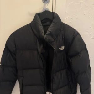 Svart pufferjacka från The North Face - Säljer en svart pufferjacka från The North Face med klassisk logga på bröstet. Jackan har hög krage, dragkedja och är quiltad för extra värme. Perfekt för kalla dagar och har en clean, oversized look som är enkel att styla.
