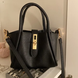 Svart handväska med gulddetaljer - Snygg svart handväska i skinnimitation med guldfärgade metalldetaljer och lås framtill. 🩷Är silver tjej och tänkte inte på att det va guld på när jag köpte så den är nu!🩷🫣 Därför jag säljer!