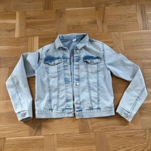 Ljusblå jeansjacka med knappar - Snygg ljusblå jeansjacka med klassisk krage och silverfärgade knappar framtill. Jackan har två bröstfickor med lock och knapp, samt långa ärmar med knapp vid mudden. Perfekt för att ge din outfit en cool touch.