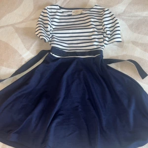 Randig marinblå klänning Holly & Whyte - Snygg kortklänning från Holly & Whyte by Lindex med vit och marinblå randig topp, rund hals och korta ärmar. Kjolen är helmarinblå och har ett matchande knytband i midjan. Perfekt för dig som gillar klassisk och fräsch stil.