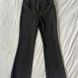 Svarta bootcut kostymbyxor Naf Naf - Snygga svarta kostymbyxor från Naf Naf i bootcut-modell med diskreta vita ränder. Byxorna har hög midja, snörningsdetaljer framtill och klassisk dragkedja med hyska och knapp. Perfekt för dig som gillar en stilren och lite edgy look. Står storlek 38 men är 36 sj och de är lite tajta hade passat xs bättre 