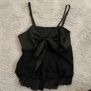 Svart linnetopp med rosett - Snygg svart linnetopp med tunna axelband och stor rosett framtill. Toppen har ett flowigt, dubbellager tyg med vågig kant nedtill som ger en cool look. Perfekt för att lyfta din outfit med lite extra stil.