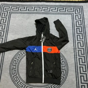 Psg jacka - Svart vindjacka från Jordan x PSG med huva, vita dragkedjor och färgblock i blått och rött över bröstet. PSG-logga och Jumpman på framsidan, stor PARIS-tryck på ryggen. Jackan har långa ärmar och två fickor med dragkedja. Perfekt för dig som gillar sportig streetstyle.