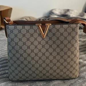 Beige axelväska med V-detalj - Snygg beige axelväska med brunt och guldiga detaljer. Väskan har ett geometriskt mönster, ett stort V i metall framtill och flera fack med dragkedja. Axelremmen är justerbar och insidan är fodrad i brunt. Perfekt för dig som gillar stilrena accessoarer.