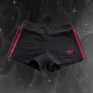 Svarta Adidas shorts med rosa ränder - Snygga svarta shorts från Adidas med ikoniska rosa ränder längs sidorna och broderad logga framtill. Modellen är tight och har bred midja med två metallöljetter framtill. Mjukt bomullsmaterial som är perfekt för träning eller chill. Klassisk streetstil med sportig vibe. S/M